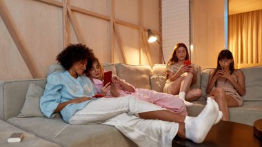 Young focused multiethnic girlfriends using smartphones on sofa during girlish sleepover at home. Black and caucasian zoomer girls rest and spend time together. Friendship. Entertainment and leisure