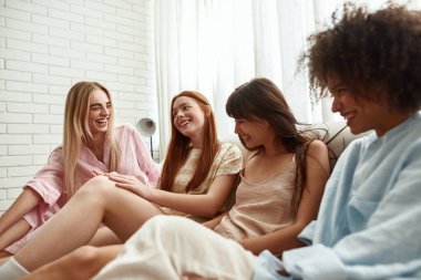 Young multiethnic girlfriends laughing and enjoying time on bed during girlish pajama party at home. Black and caucasian zoomer girls resting together. Friendship. Bedroom in modern apartment