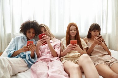 Young multiracial girlfriends using and watching smartphones on bed during girlish pajama party at home. Black and caucasian zoomer girls spending time together. Friendship. Entertainment and leisure
