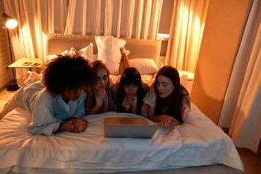 Young concentrated multiethnic girlfriends watching laptop on bed during girlish sleepover. Black and caucasian zoomer girls resting and spending time together. Friendship. Entertainment and leisure