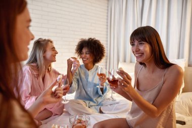 Young multiethnic girlfriends drinking wine and feeding marshmallow each other on bed during girlish sleepover. Black and caucasian zoomer girls enjoying time together. Friendship. Rest and leisure