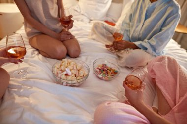 Cropped image of girlfriends drinking wine and eating dessert snacks on bed during girlish sleepover at night home. Zoomer girls rest and spend time together. Friendship. Entertainment and leisure