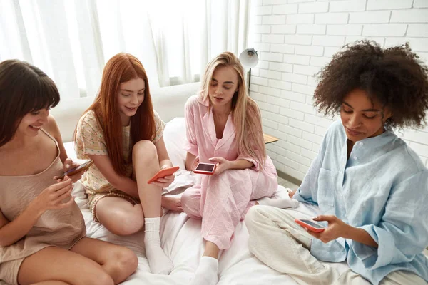 Young concentrated multiracial girlfriends using and browsing smartphones on bed during girlish pajama party at home. Black and caucasian zoomer girls. Friendship. Rest, entertainment and leisure