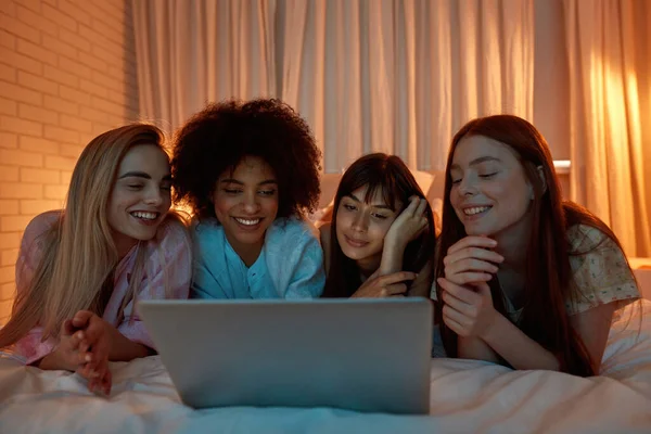 Young multiracial girlfriends watching laptop on bed during girlish sleepover. Black and caucasian zoomer girls spending time together. Friendship. Entertainment and leisure. Evening on night time