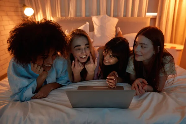 Young cheerful multiracial girlfriends enjoying while watching laptop on bed during girlish sleepover at night. Black and caucasian zoomer girls resting together. Friendship. Entertainment and leisure