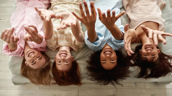 Top view of young smiling multiracial girlfriends lying on bed and pulling hands up during girlish pajama party at home. Black and caucasian zoomer girls resting and spending time together. Friendship
