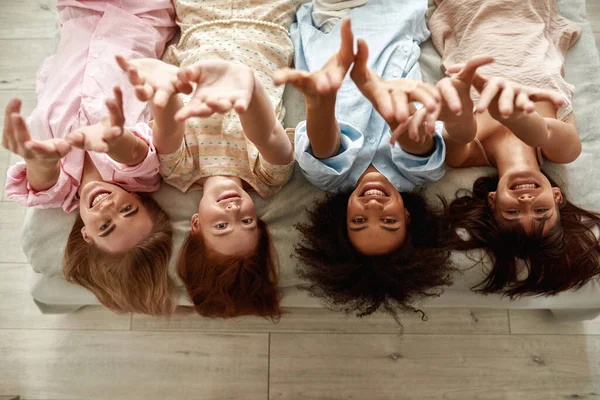 Top view of four young smiling multiethnic girlfriends lying on bed and pulling hands up during girlish pajama party at home. Black and caucasian zoomer girls rest and spend time together. Friendship