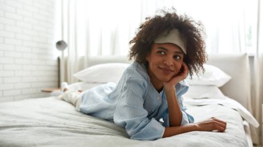 Young smiling black girl lying and looking away on bed at home in morning. Beautiful curly teen female zoomer wearing homewear and sleep mask . Domestic lifestyle. Modern bedroom in spacious apartment