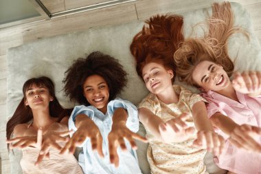 Top view of young smiling multiracial girlfriends lying on bed and pulling hands up during girlish pajama party at home. Black and caucasian zoomer girls resting and looking at camera. Friendship