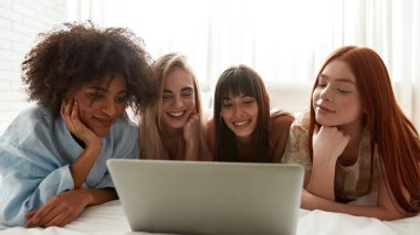 Young positive multiethnic girlfriends watching laptop on bed during girlish pajama party. Black and caucasian zoomer girls resting and enjoying time together. Friendship. Entertainment and leisure