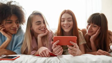 Selective focus on smartphone in hand of red haired girl show something to multiracial girlfriends on bed at girlish pajama party at home. Smiling girls. Friendship. Rest, entertainment and leisure