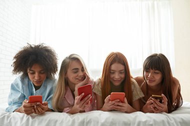 Young multiracial girlfriends chatting and messaging on smartphones on bed during girlish pajama party at home. Black and caucasian zoomer girls resting together. Friendship. Entertainment and leisure