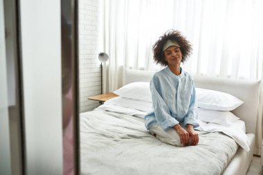 Young black girl sitting and looking at camera on bed at home in morning. Beautiful curly teen female zoomer wearing homewear and sleep mask. Domestic lifestyle. Modern bedroom in spacious apartment