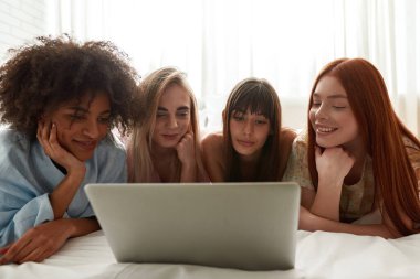 Young focused multiracial girlfriends watching laptop on bed during girlish pajama party. Black and caucasian zoomer girls resting and enjoying time together. Friendship. Entertainment and leisure
