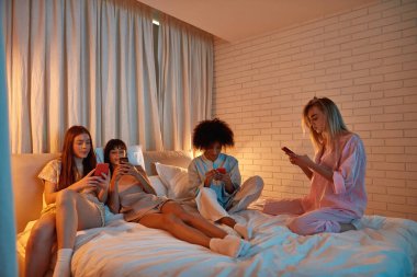 Young focused multiracial girlfriends use smartphones on bed during girlish sleepover. Black and european zoomer girls spend time together. Friendship. Entertainment and leisure. Evening or night time