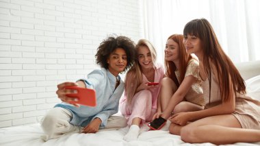 Young smiling multiethnic girlfriends take selfie on smartphone on bed during girlish pajama party at home. Black and caucasian zoomer girls enjoy time together. Friendship. Entertainment and leisure