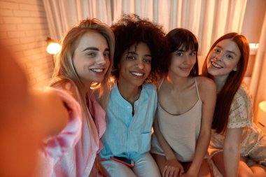 Young smiling multiethnic girlfriends looking at camera while taking photo or selfie during girlish sleepover at night time. Black and caucasian girls. Friendship. Rest, entertainment and leisure