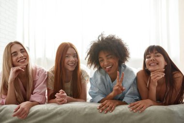 Young smiling multiracial girlfriends lying on bed and looking at camera during girlish pajama party. Black girl winking, showing victory sign and sticking out tongue. Friendship. Modern bedroom