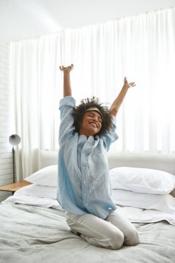Young smiling black girl waking up on bed at home in morning time. Curly teen female zoomer with closed eyes wearing homewear and sleep mask. Domestic lifestyle. Modern bedroom in spacious apartment