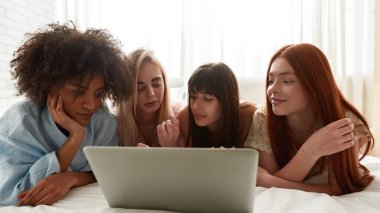 Young multiethnic girlfriends lying on bed and watching laptop during girlish pajama party at home. Black and caucasian zoomer girls rest and spend time together. Friendship. Entertainment and leisure