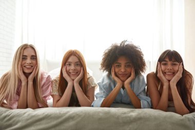 Young smiling multiracial girlfriends lying on bed and looking at camera during girlish pajama party. Black and caucasian zoomer girls resting and spending time together. Friendship. Modern bedroom
