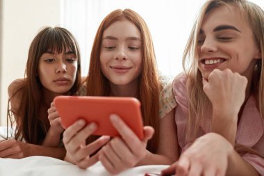 Young caucasian girlfriends watching smartphone in hand of red haired girl on bed during girlish pajama party at home. Zoomer girls spending time together. Friendship. Rest, entertainment and leisure