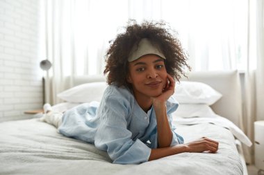 Young smiling black girl lying and resting on bed at home in morning time. Pretty curly teen female zoomer wearing homewear and sleep mask looking at camera. Domestic lifestyle. Spacious bedroom