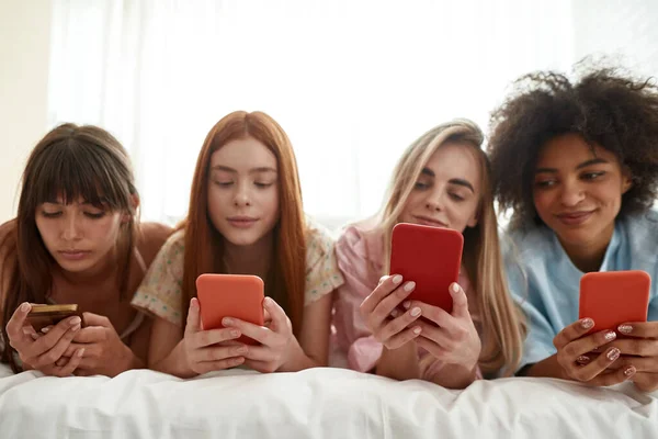 Young multiracial girlfriends use and watch smartphones on bed during girlish pajama party at home. Black and caucasian zoomer girls spending time together. Friendship. Rest, entertainment and leisure
