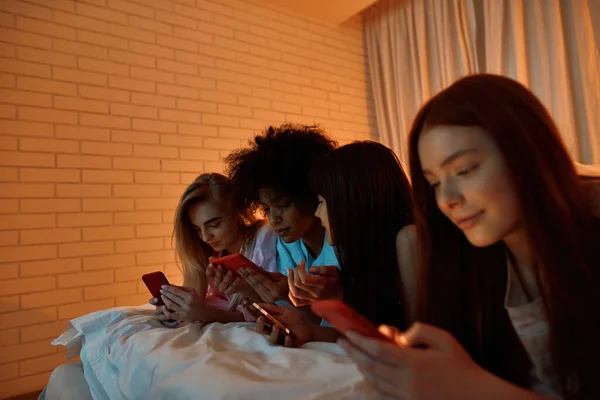 Young multiethnic girlfriends use and watch smartphones on bed during girlish sleepover at home. Black and caucasian zoomer girls rest and spend time together. Friendship. Entertainment and leisure