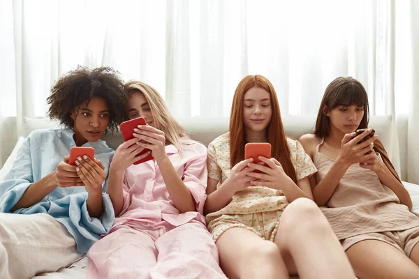 Young multiethnic girlfriends browsing smartphones on bed during girlish pajama party at home. Black and european zoomer girls resting and spending time together. Friendship. Entertainment and leisure