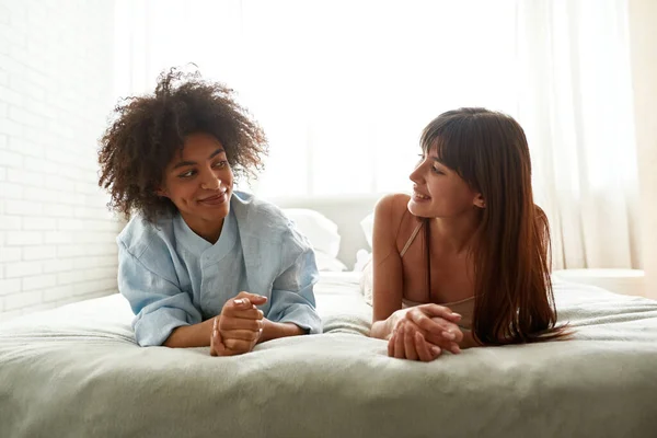 Young Smiling Multiethnic Girlfriends Lying Bed Looking Camera Home Black — Zdjęcie stockowe