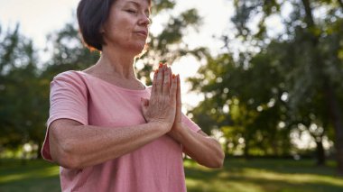 Partial of focused caucasian senior woman with closed eyes practicing yoga and meditating in blurred green park. Sportive female pensioner wearing pink t-shirt. Concept of mental health. Sunny day