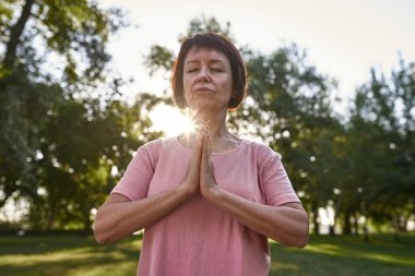 Concentrated caucasian mature woman with closed eyes practicing yoga and meditating in blurred green park. Sportive female pensioner wearing pink t-shirt. Concept of healthy lifestyle. Sunny day