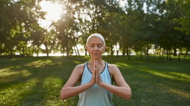 Caucasian senior woman with closed eyes practicing yoga and meditating on green meadow in park. Sportive female pensioner wearing sportswear. Concept of healthy lifestyle. Sunny day