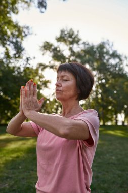 Focused caucasian senior woman with closed eyes practicing yoga and meditating on green meadow in park. Sportive female pensioner wearing pink t-shirt. Concept of healthy lifestyle. Sunny day