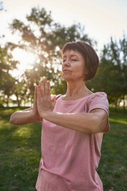 Concentrated european mature woman with closed eyes practicing yoga and meditating on green lawn in park. Sportive female pensioner wearing pink t-shirt. Concept of healthy lifestyle. Sunny summer day