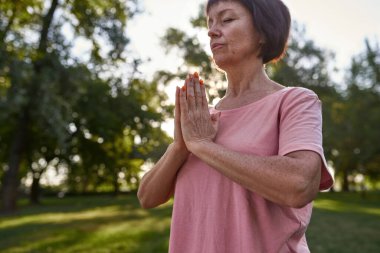 Cropped of concentrated caucasian middle aged woman with closed eyes practicing yoga and meditating in blurred green park. Sportive female pensioner wearing pink t-shirt. Healthy lifestyle. Sunny day
