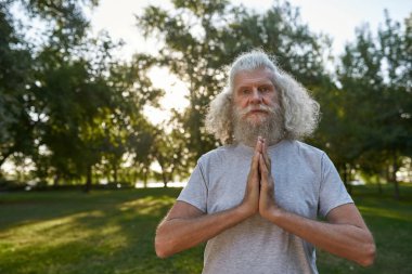 Serious caucasian elderly man practicing yoga and meditating on green meadow in park. Sportive bearded male pensioner wearing sportswear looking at camera. Concept of healthy lifestyle. Warm sunny day