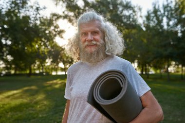 Partial of smiling caucasian elderly man with fitness mat looking at camera on green meadow in park. Sportive bearded male pensioner wearing sportswear. Concept of healthy lifestyle. Sunny summer day