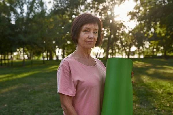 Serious senior caucasian woman with fitness mat on green meadow in park. Sportive female pensioner wearing sportswear looking at camera. Concept of healthy lifestyle. Warm sunny day