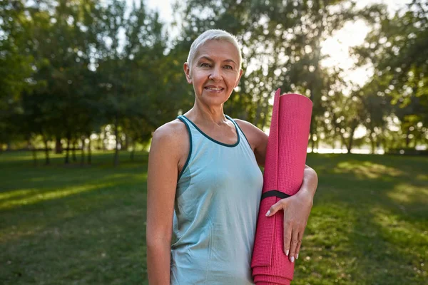Smiling caucasian senior woman with fitness mat on green meadow in park. Sportive female pensioner wearing sportswear looking at camera. Concept of healthy lifestyle. Warm sunny day