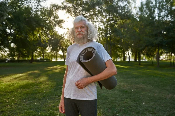 Serious caucasian elderly man with fitness mat on green lawn in park. Sportive bearded male pensioner wearing sportswear looking at camera. Concept of healthy lifestyle. Summer sunny day