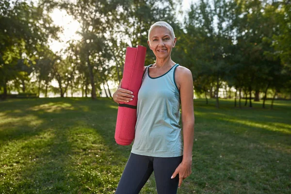Smiling caucasian middle aged woman with fitness mat on green meadow in park. Sportive female pensioner wearing sportswear looking at camera. Concept of healthy lifestyle. Warm sunny day