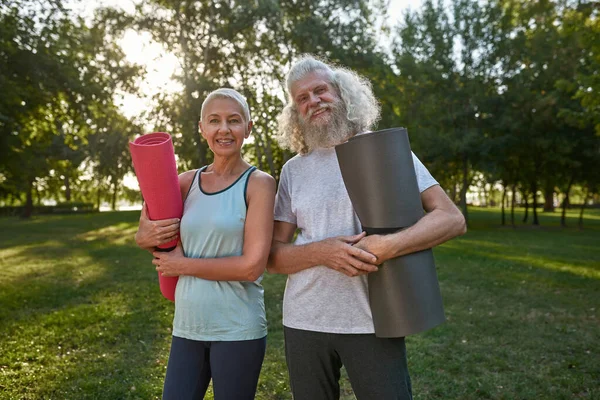 Smiling caucasian elderly couple with fitness mats on green lawn in park. Sportive man and woman wearing sportswear looking at camera. Concept of healthy lifestyle. Sunny summer day