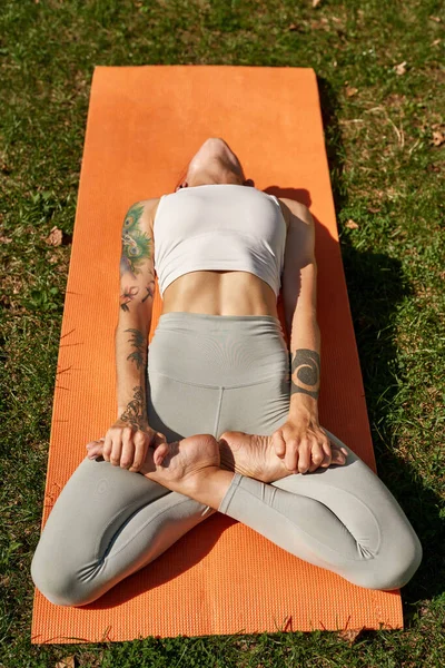 Top view of young sportswoman practicing yoga in fish pose in sunny park. Beautiful fit girl with tattoos wearing sportswear on fitness mat on green meadow. Concept of healthy lifestyle
