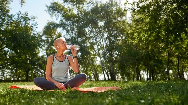 European elderly woman drinking water and resting after sports training in green sunny park. Sportive female with closed eyes wearing sportswear sitting on fitness mat on lawn. Healthy lifestyle