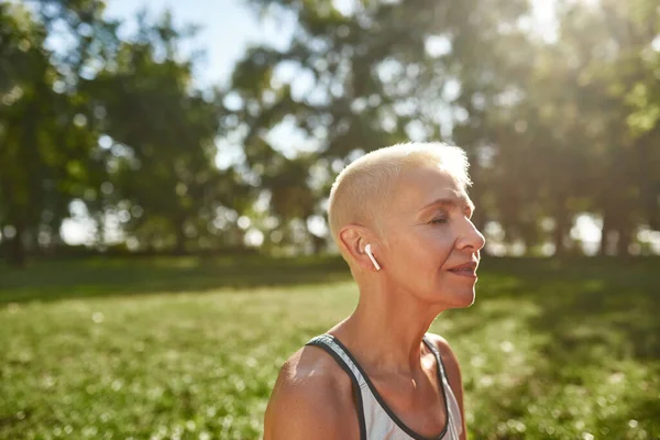 Partial of caucasian grey hair woman practicing yoga and meditating on green lawn in sunny park. Sportive female pensioner with closed eyes wearing sportswear. Concept of healthy lifestyle