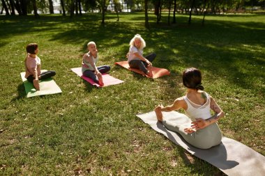 Female trainer teaching elderly man and women how practicing yoga in half bound lotus forward fold pose in sunny park. Sportive people on fitness mats on green lawn. Concept of healthy lifestyle