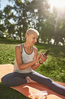Smiling caucasian elderly woman browsing smartphone in sunny park. Sportive female pensioner wearing sportswear sitting on fitness mat on green meadow. Concept of healthy lifestyle