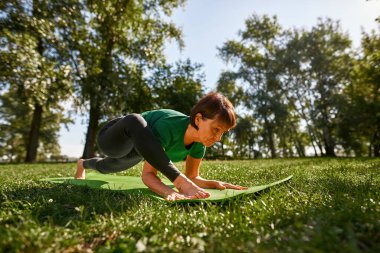 Concentrated caucasian elderly woman practicing yoga in sunny park. Sportive female pensioner wearing sportswear standing on fitness mat on green meadow. Concept of healthy lifestyle